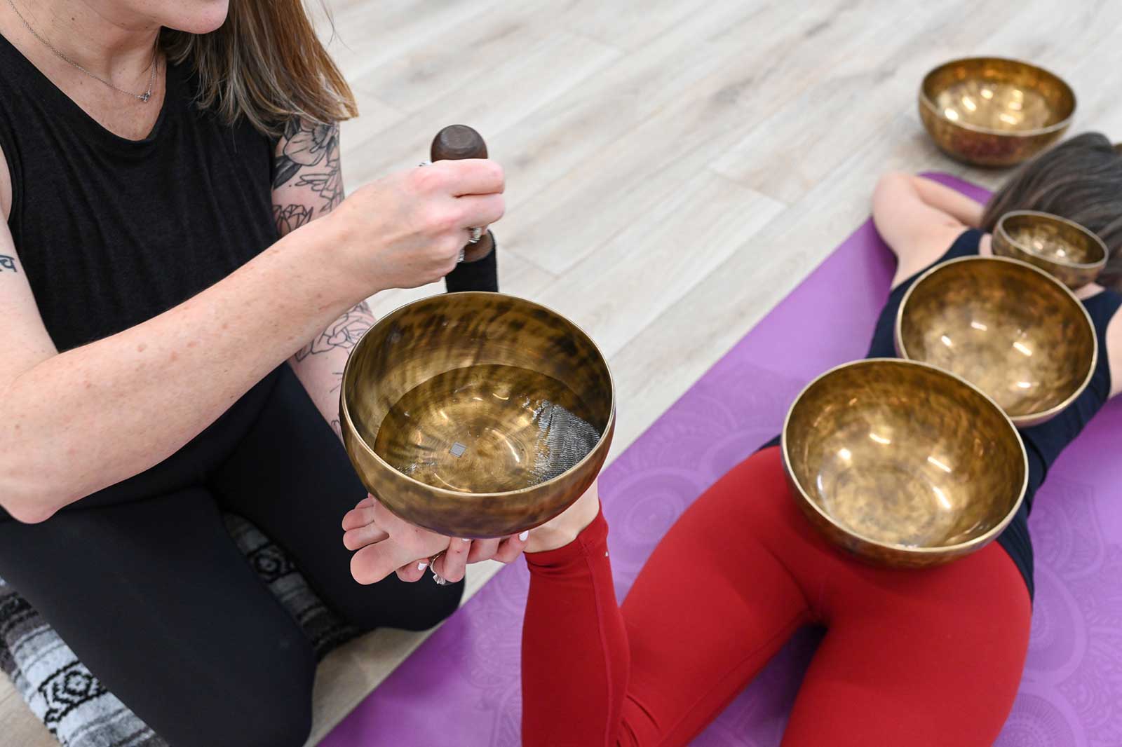 Michelle Young wearing a black yoga top and bottoms seated in front of a woman in black top and red bottoms lying face down on a purple yoga mat with 3 brass singing bowls balanced on her back body as Michelle Young holds a singing bowl half full of water on the students upward facing foot
