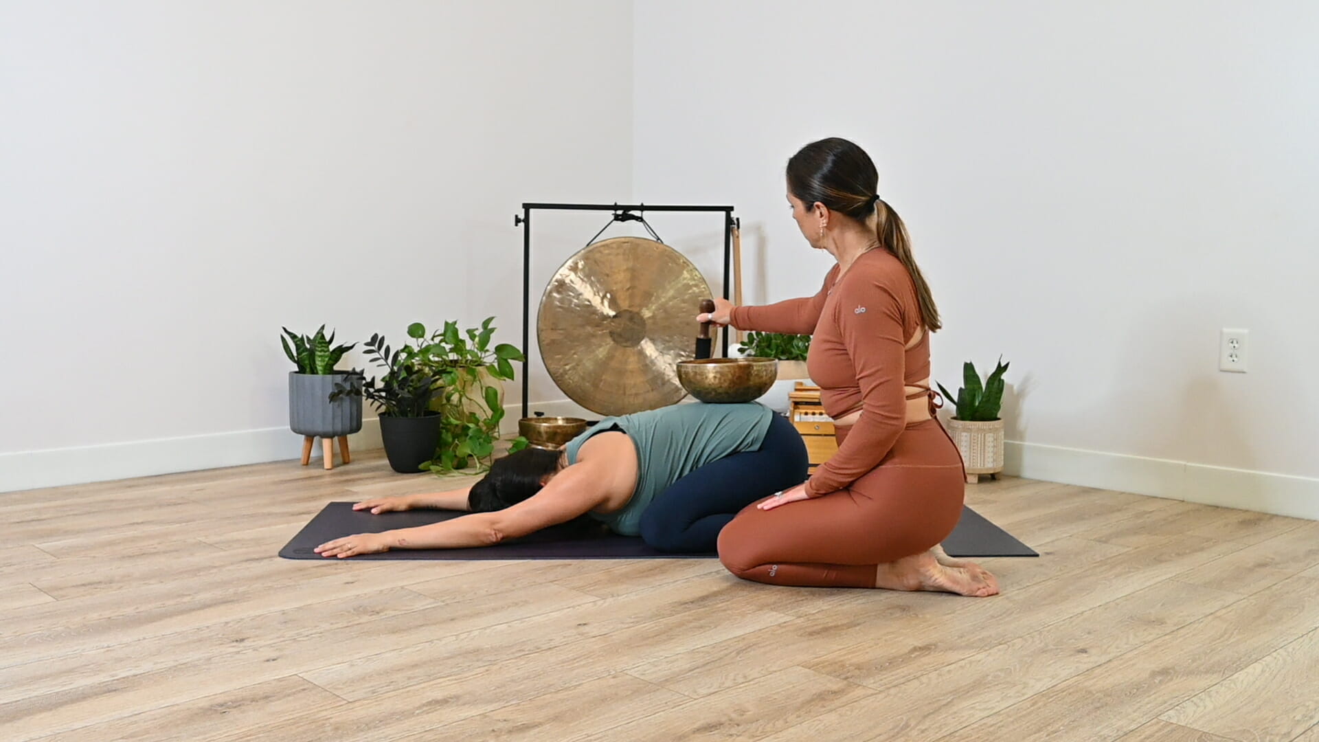 Woman in child's pose with brass bowl on her back while teacher plays bras bowl softly.