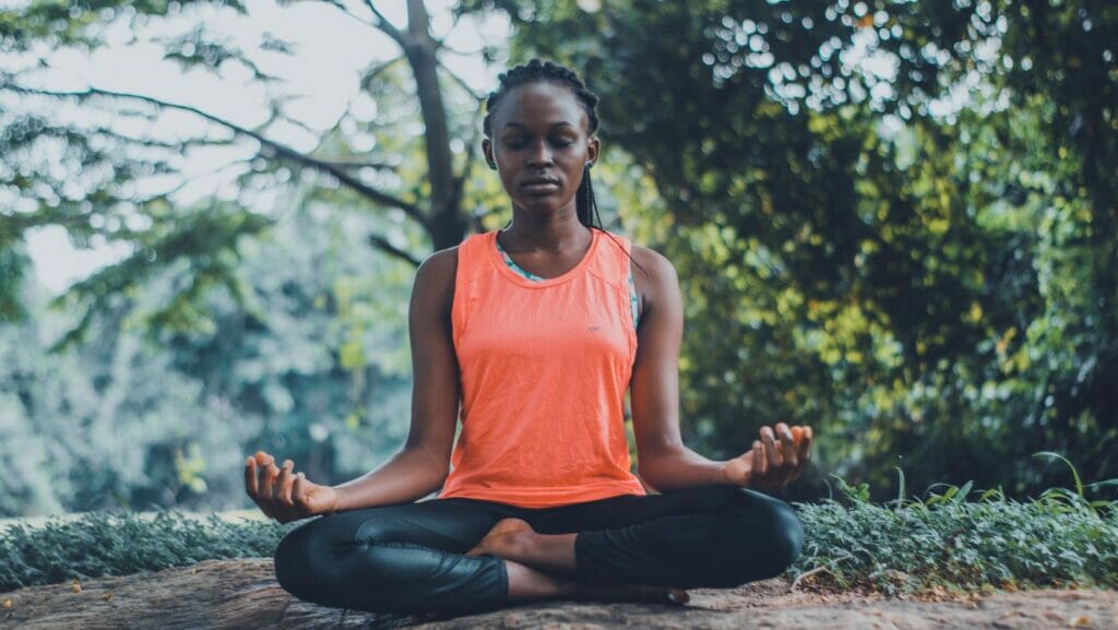 A woman with black hair sits on the ground in surrounded by trees wearing an orange shirt and black yoga plants.