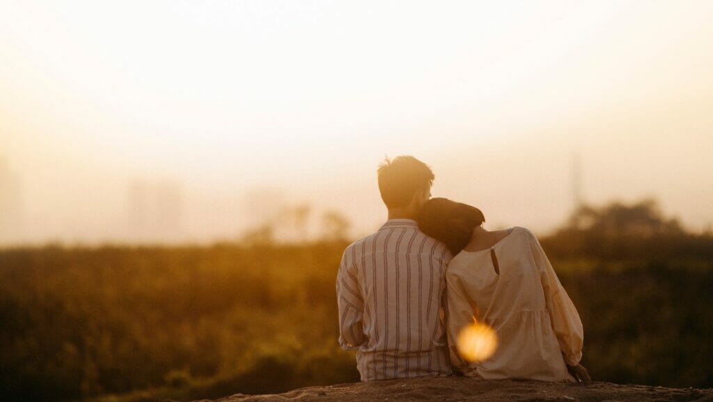 Two people sit together with backs facing the camera on a hill looking out over a field of grass. One of them leans their head on the others' shoulder.