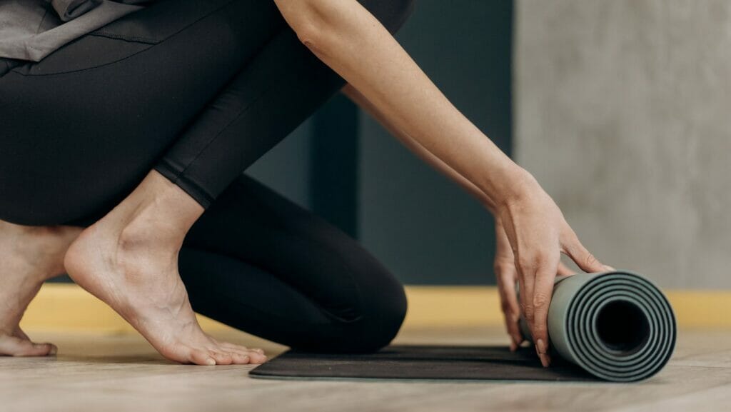 Someone in black yoga pants squats to roll up their light green yoga mat.