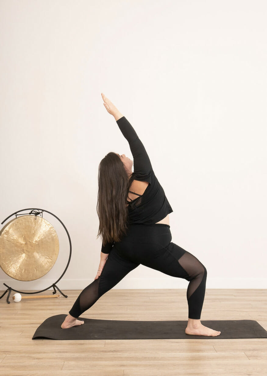 A woman in a yoga class standing on a yoga mat. She is in a reverse-warrior yoga pose.