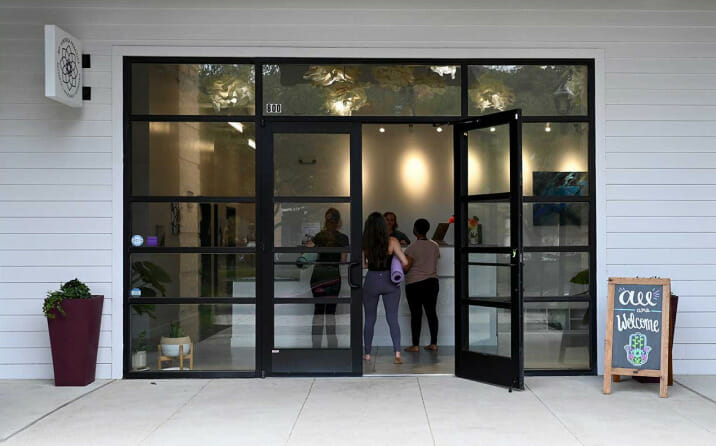The MVP studio which has large windows in the front of the studio. The windows look into a bright and welcoming storefront. There is a sign that says "all are welcome". There are women inside, holding yoga mats and going to a yoga class.