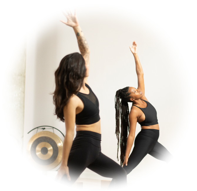 Two women yogis are in a yoga class. Their right arms are held straight up in the air.